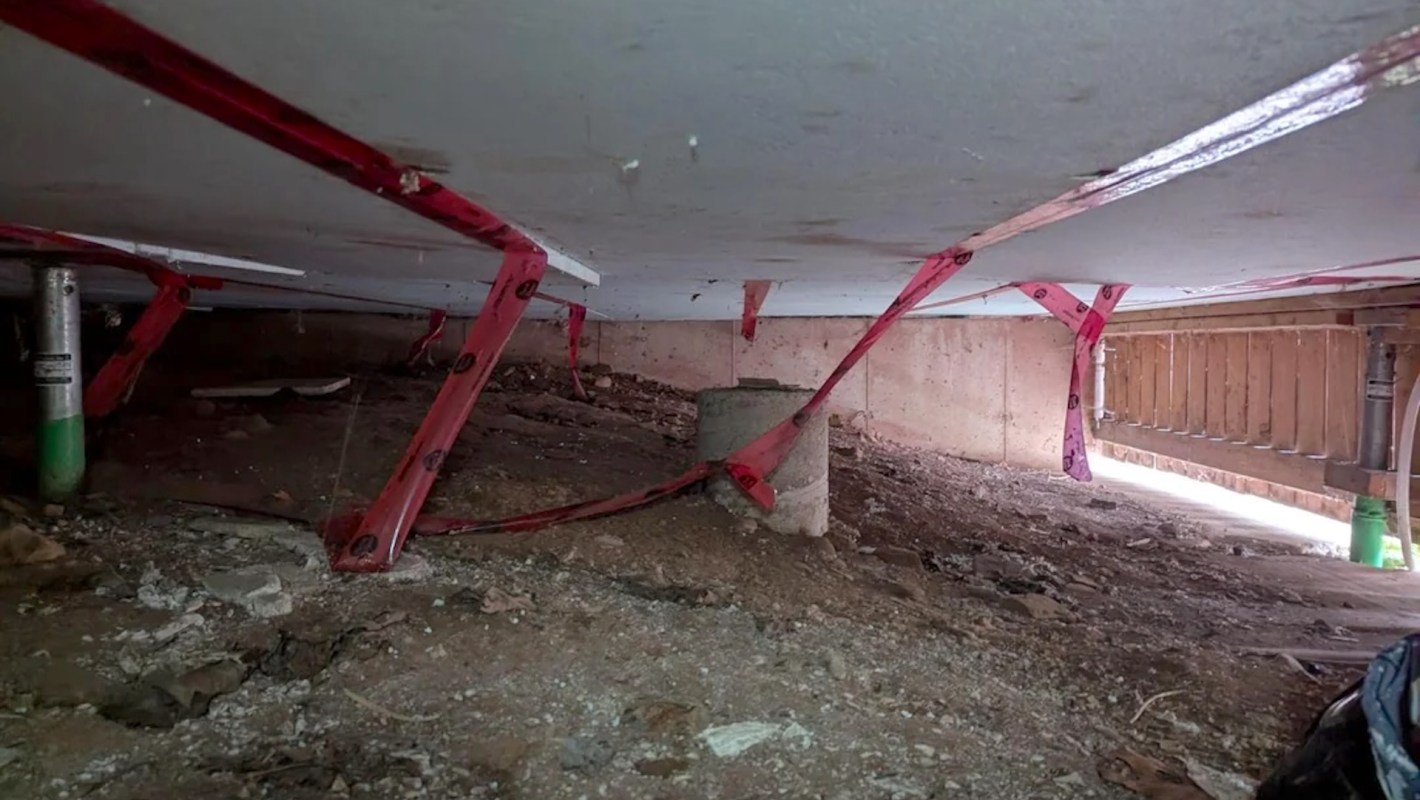 A view of the underside of a structure with red support straps and concrete foundations among loose soil.
