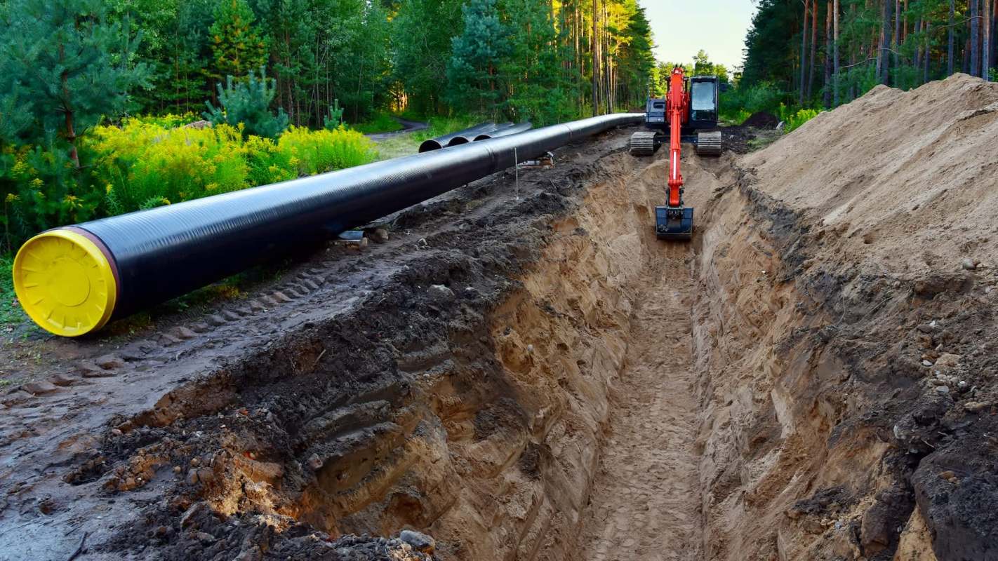 An excavator works beside a large black gas pipe along a trench in a forested area.