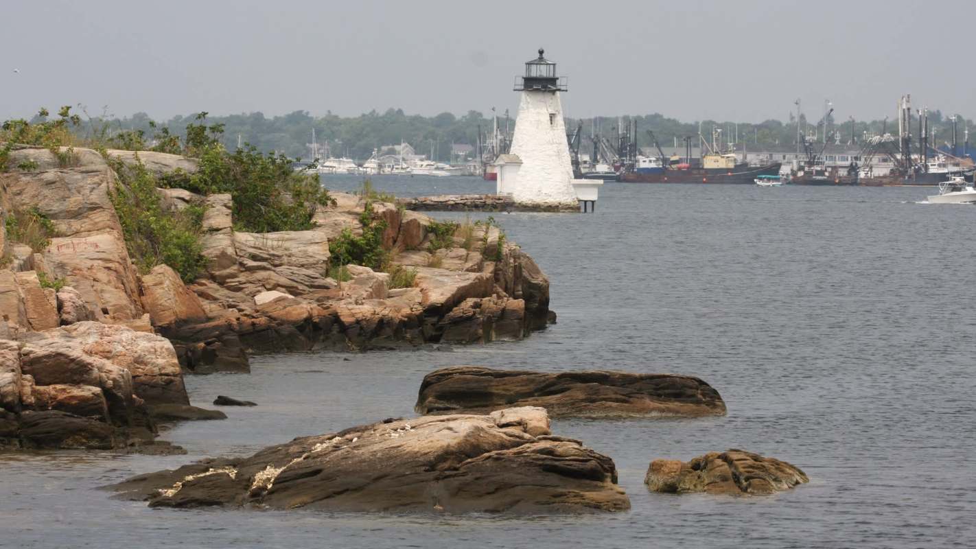 A lighthouse stands on a rocky shoreline with boats in the distance on a calm waterway.