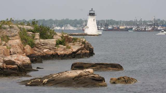 A lighthouse stands on a rocky shoreline with boats in the distance on a calm waterway.