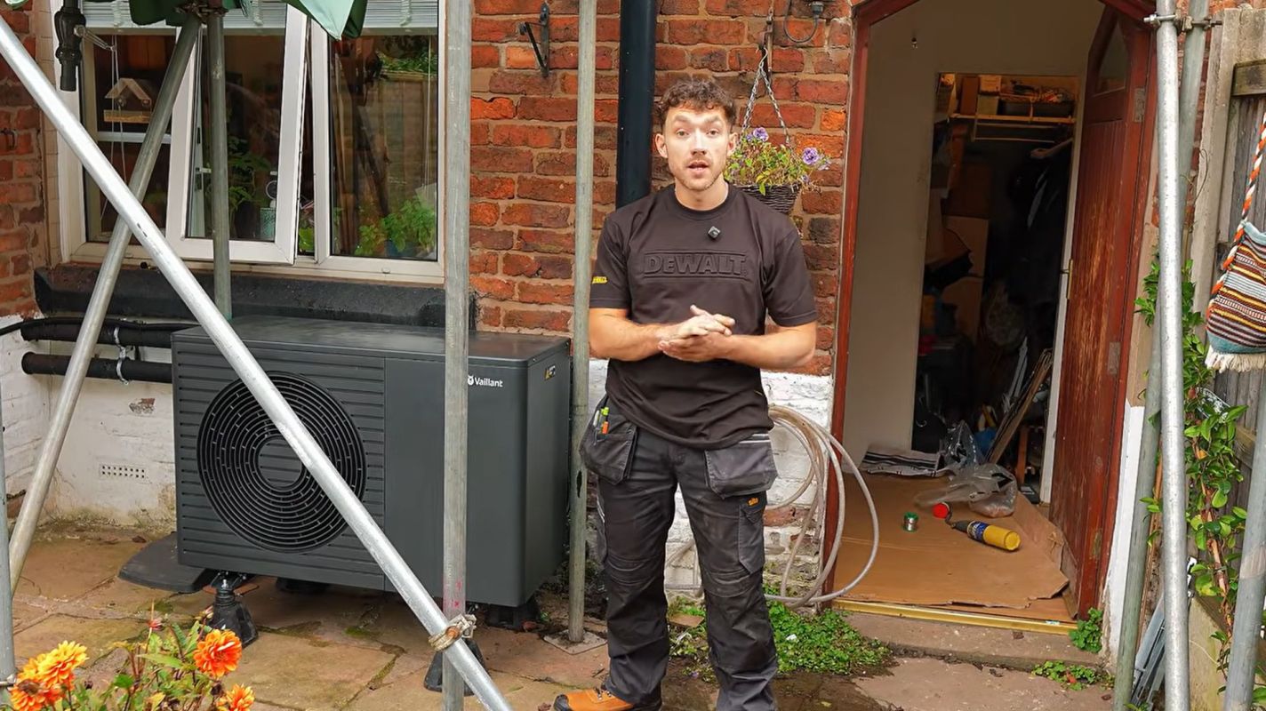 A person stands near an outdoor air conditioning unit in a residential setting with gardening elements visible.