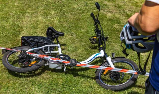 An electric bike secured with caution tape on grass, with a helmet held by a person nearby.