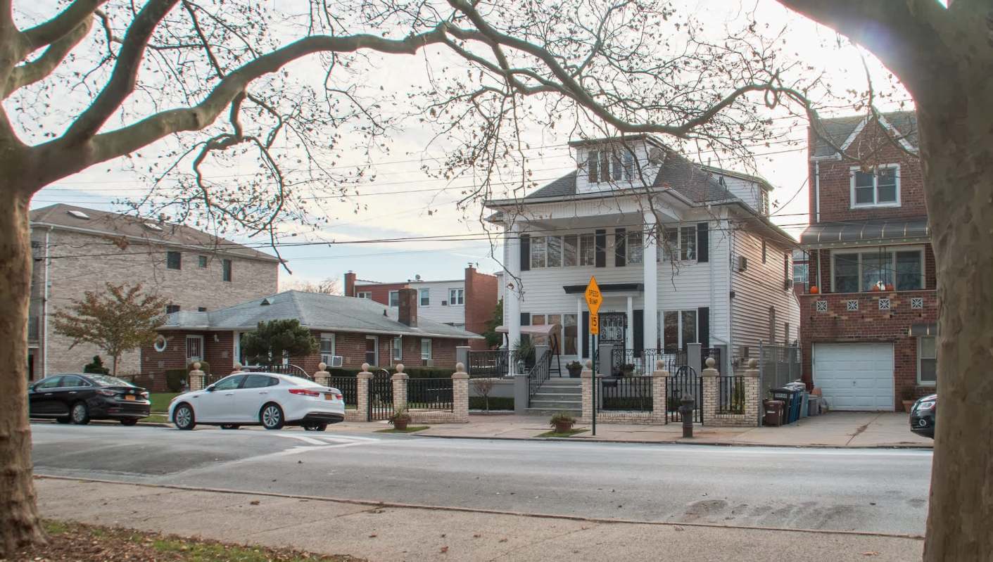 A two-story house with a porch, surrounded by a fence, next to other residential buildings and parked cars.