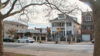A two-story house with a porch, surrounded by a fence, next to other residential buildings and parked cars.