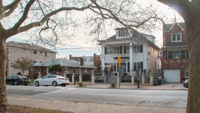 A two-story house with a porch, surrounded by a fence, next to other residential buildings and parked cars.