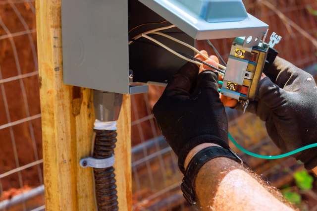 A person working on electrical wiring inside a gray electrical box mounted on a wooden post.