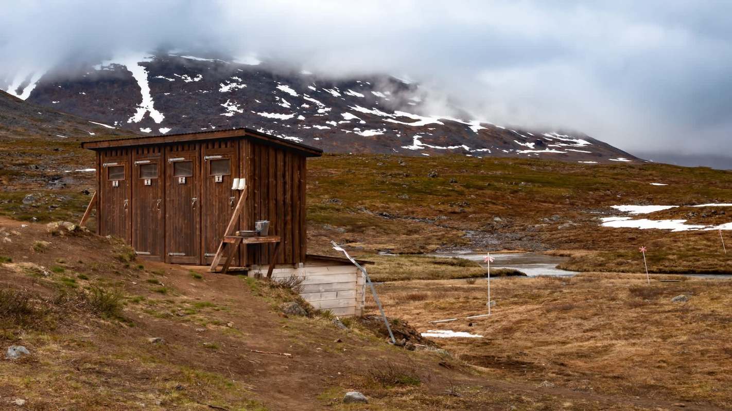 A wooden structure with multiple doors near a grassy area and a stream, surrounded by mountains and clouds.