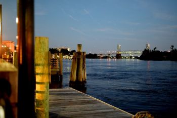 A wooden dock extends over calm water with a bridge and city lights in the background at twilight.