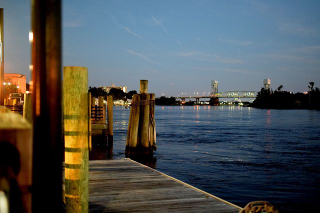A wooden dock extends over calm water with a bridge and city lights in the background at twilight.