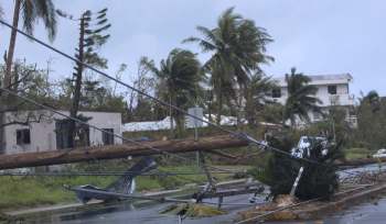 A street scene with fallen power lines, damaged buildings, and palm trees in a storm-affected area.