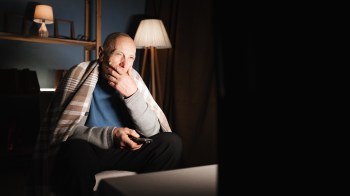 An older man wrapped in a blanket watches television while holding a remote.