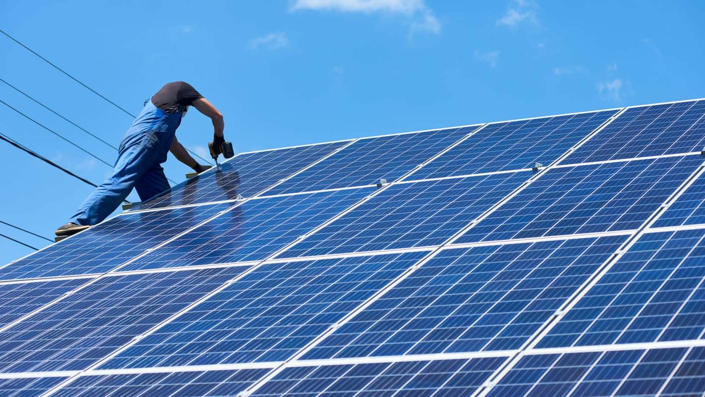 A person installs solar panels on a roof under a clear blue sky.