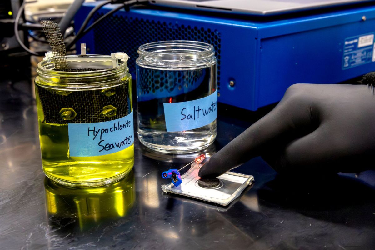 A person in a black glove touches a device near jars labeled "Hypochlorite Seawater" and "Saltwater."