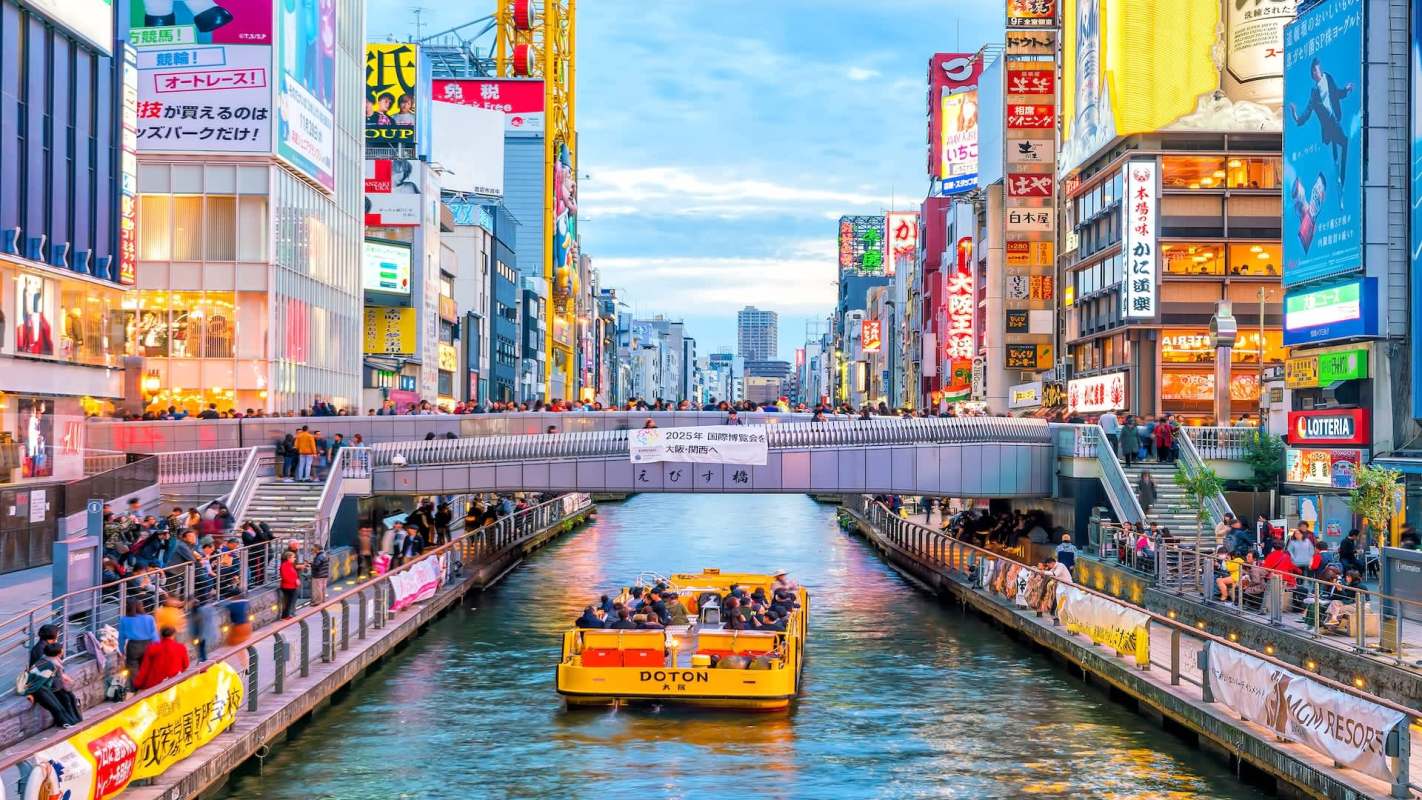 A yellow boat travels along a waterway lined with shops and illuminated signs in an urban setting.