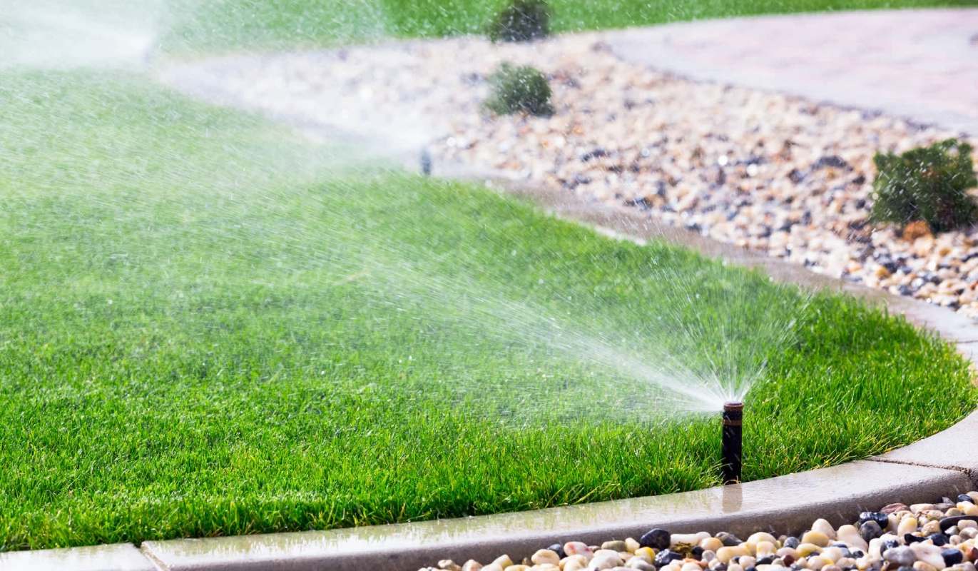A sprinkler waters a green lawn bordered by pebbles and paved pathways.