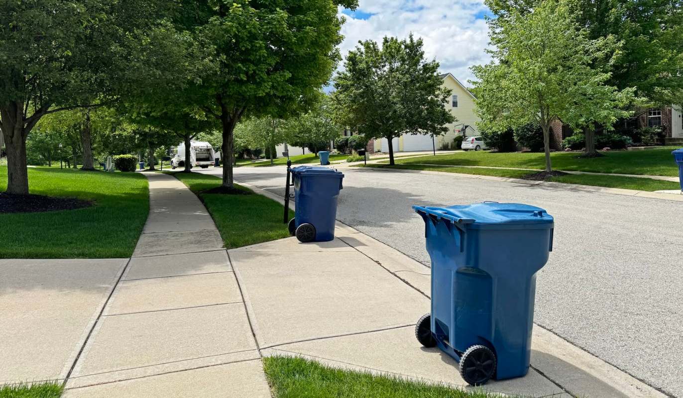 Two blue trash bins are placed along a sidewalk in a suburban neighborhood with trees and houses in the background.
