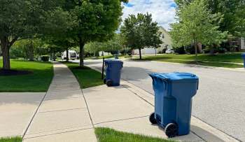 Two blue trash bins are placed along a sidewalk in a suburban neighborhood with trees and houses in the background.