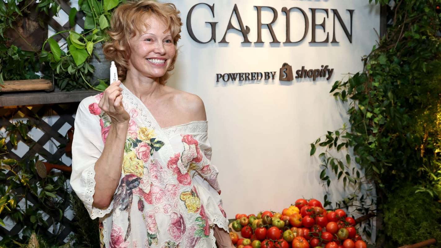 Pamela Anderson holds a marker in front of a sign that says "GARDEN" and displays a variety of tomatoes.