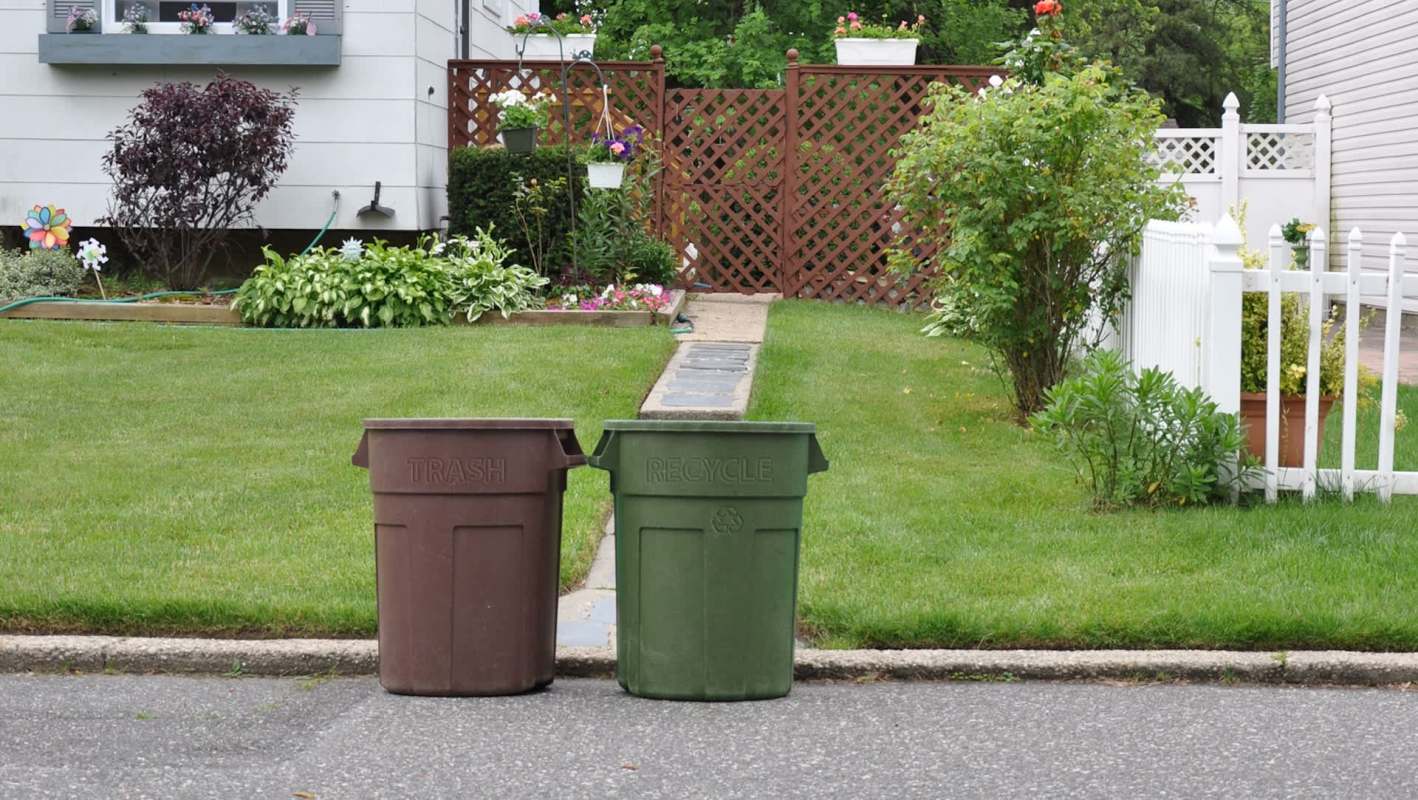 Two trash bins labeled "TRASH" and "RECYCLE" placed on a green lawn beside a garden pathway and a white fence.