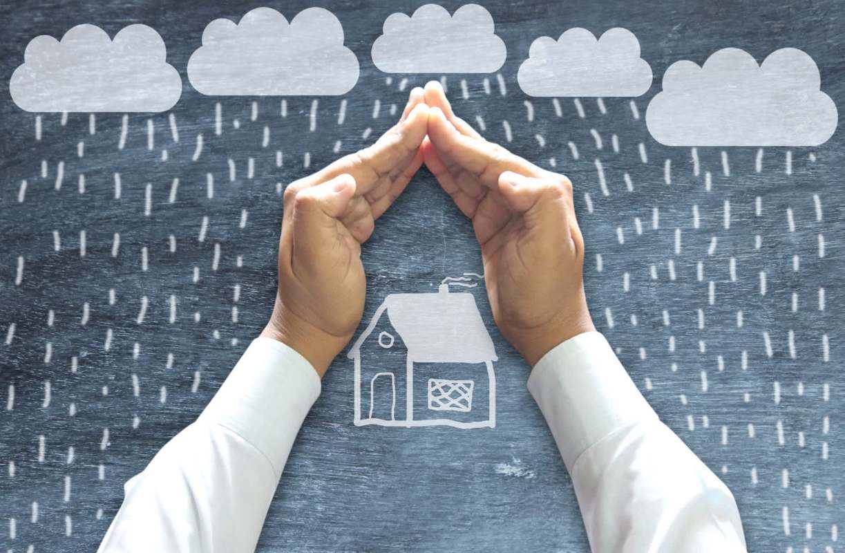 Hands forming a roof over a chalkboard drawing of a house in a storm.