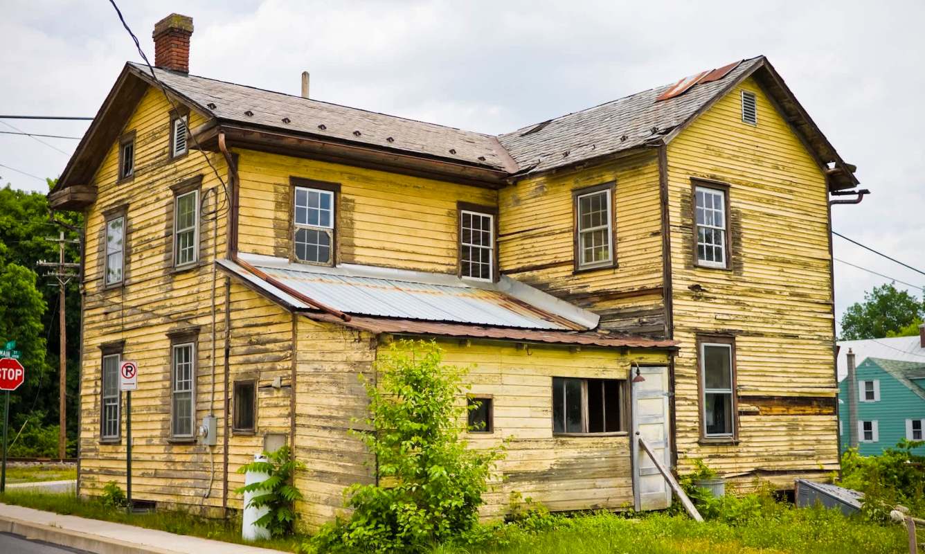 An old, weathered yellow house with multiple windows and a metal roof, surrounded by greenery.
