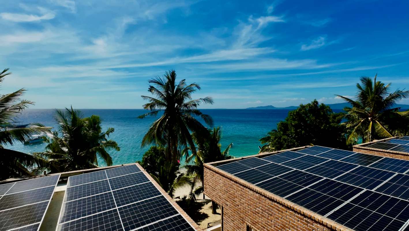 Solar panels on a building with a view of palm trees and the ocean under a blue sky.
