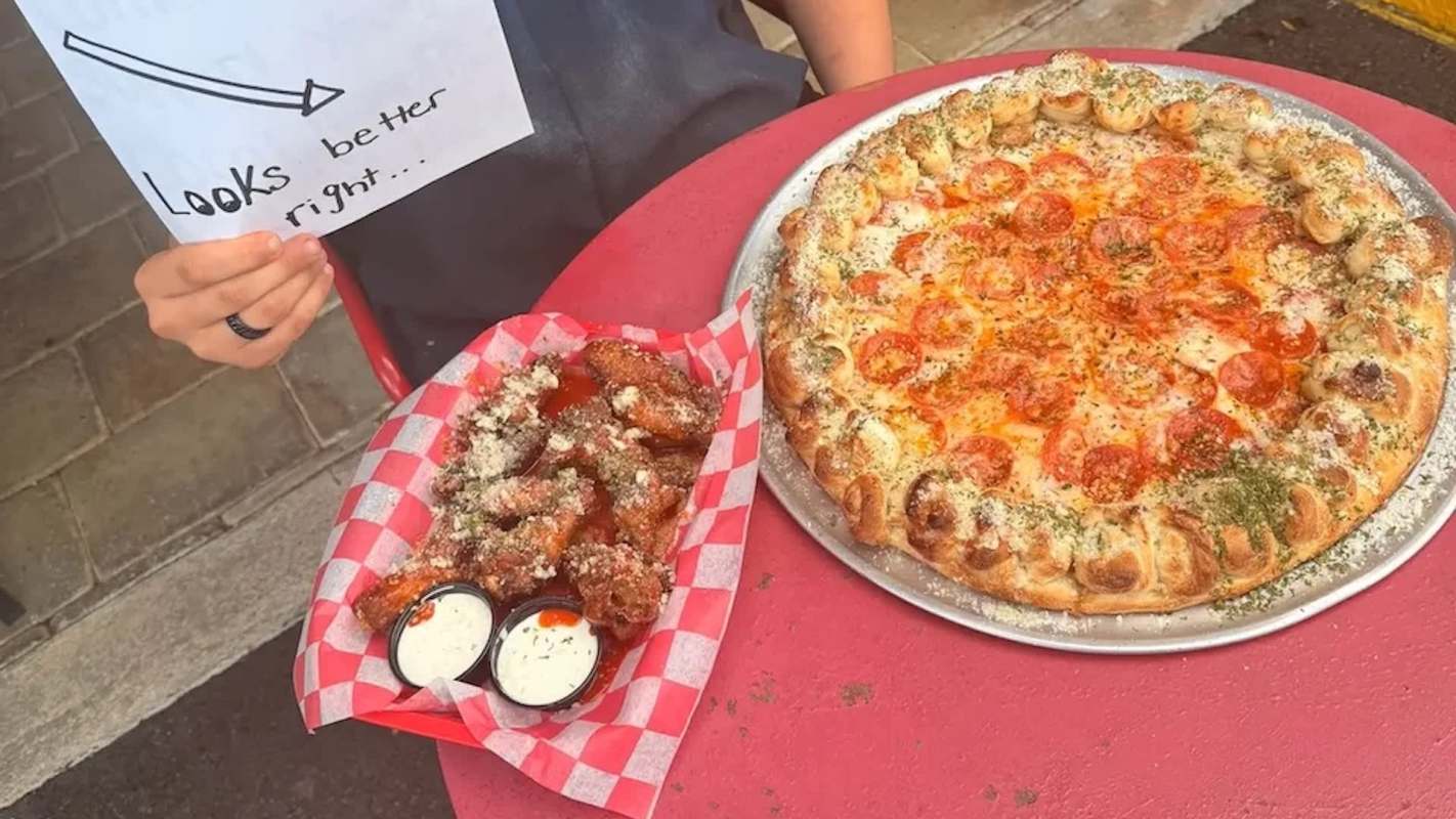 A pizza with pepperoni and a basket of chicken wings with dipping sauces on a red table.