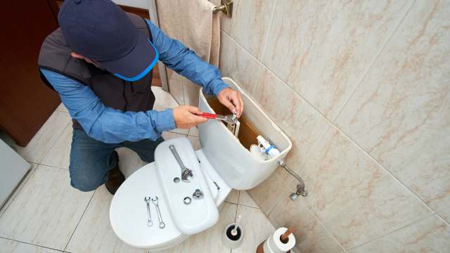 A person repairs the inside of a toilet tank using a wrench, with tools and a toilet brush nearby.