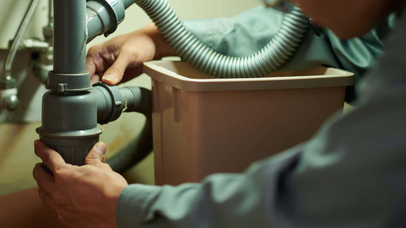 A person working on plumbing under a sink, adjusting pipes and hoses connected to a container.
