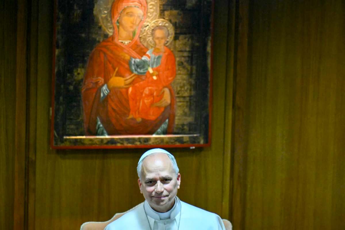 A man in a white clerical outfit sits in front of a religious icon.