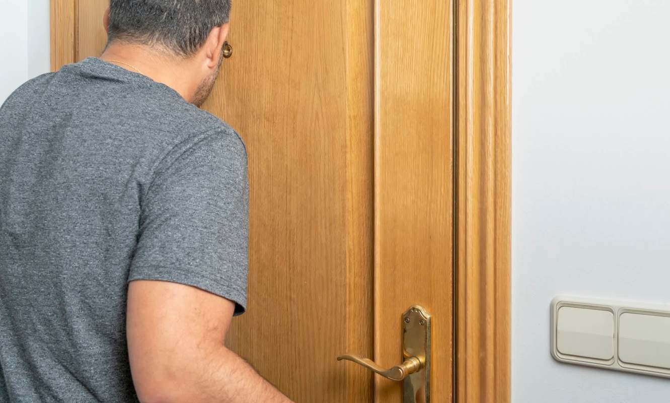 A person leaning toward a wooden door, looking through a peephole.