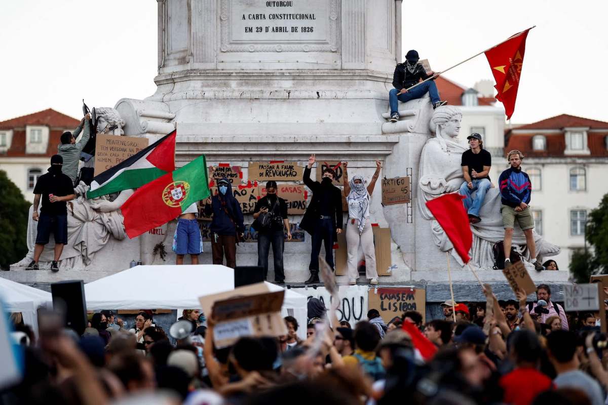 A crowd gathers around a monument in Portugal, with people holding flags and signs during a protest.