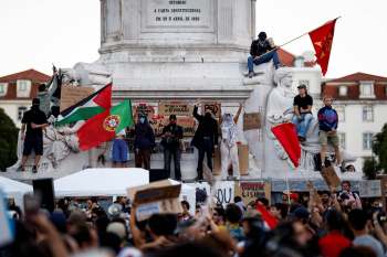 A crowd gathers around a monument in Portugal, with people holding flags and signs during a protest.