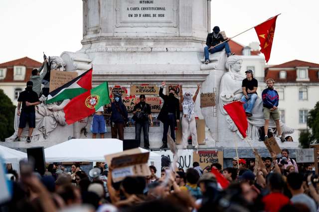 A crowd gathers around a monument in Portugal, with people holding flags and signs during a protest.