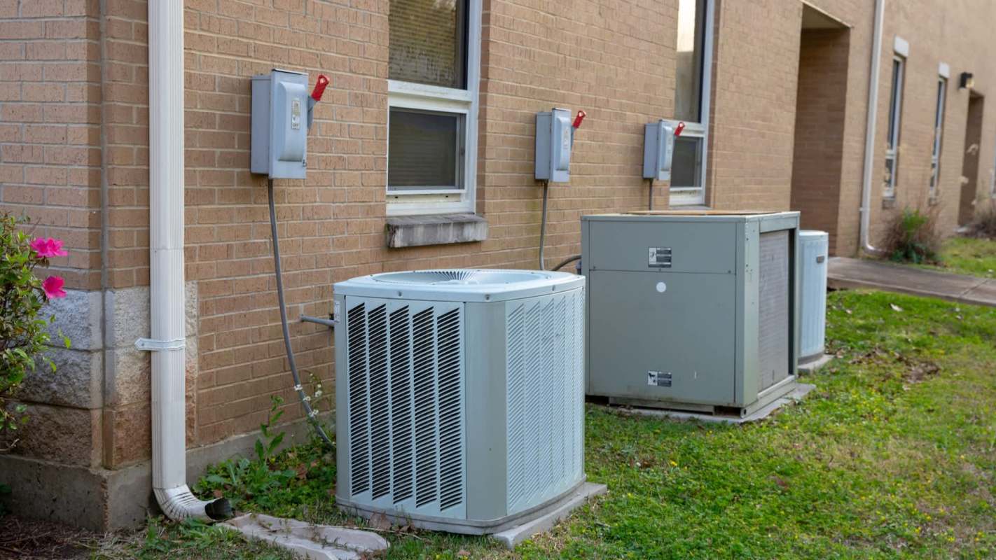 Air conditioning units and electrical boxes are installed next to a brick building, with greenery nearby.