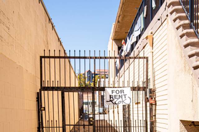 A black metal gate with a "For Rent" sign in a narrow alley between two buildings.