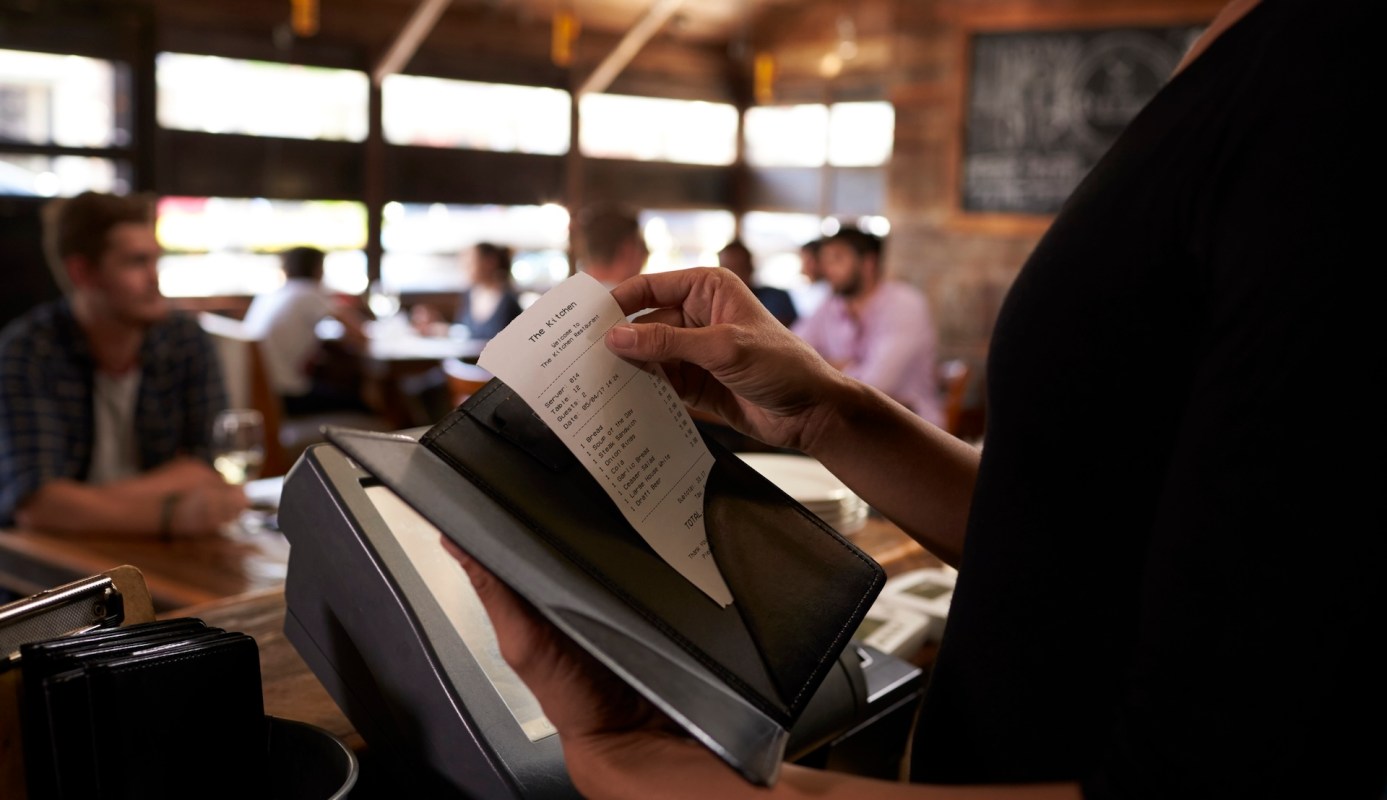 A person holds a receipt in a restaurant with diners seated in the background.