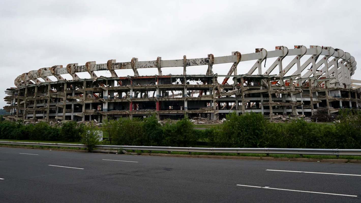 A partially collapsed stadium with visible structural damage and overgrown vegetation nearby.