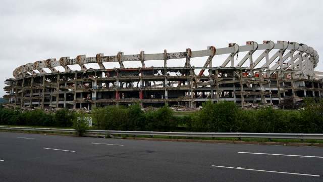 A partially collapsed stadium with visible structural damage and overgrown vegetation nearby.
