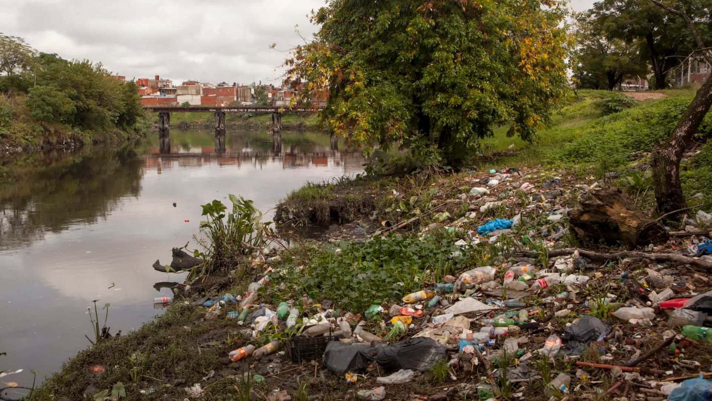 A polluted riverbank in Buenos Aires, Argentina, covered in litter and surrounded by plants and trees.