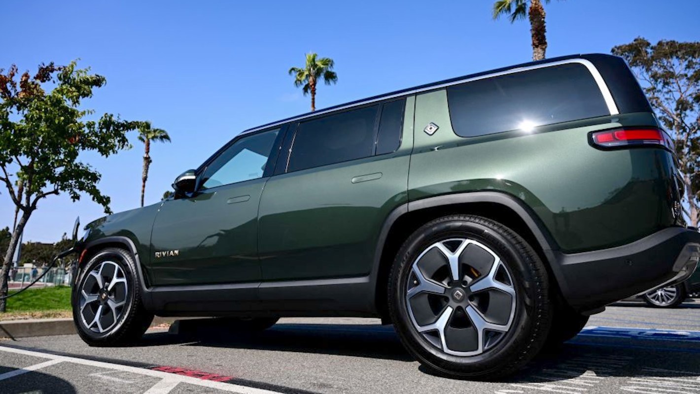 A green electric Rivian R1S SUV parked in a lot near palm trees and clear blue sky.