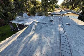A view of several roofs with grey shingles, surrounded by trees and a green lawn.