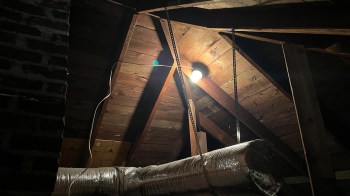An attic with exposed wooden beams, a bulb hanging, and insulated ductwork.