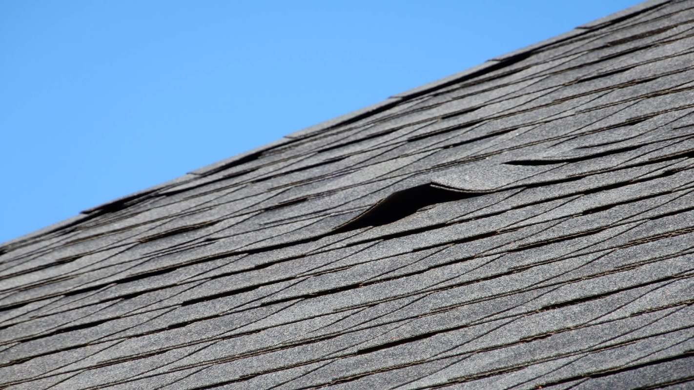 A roof with damaged and lifted shingles against a clear blue sky.