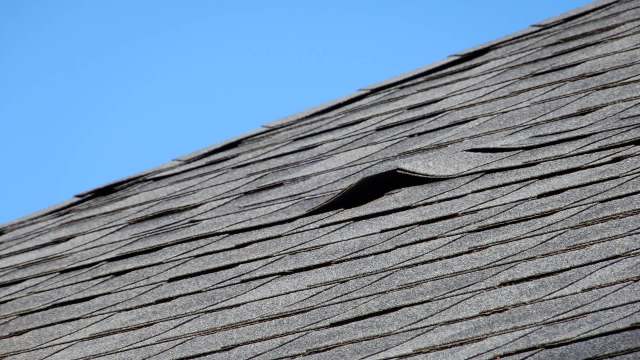 A roof with damaged and lifted shingles against a clear blue sky.