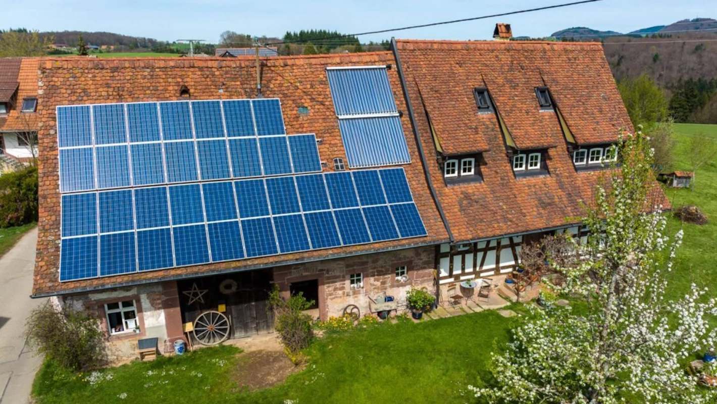 A house with a tiled roof features solar panels on one side, surrounded by a grassy area and trees.