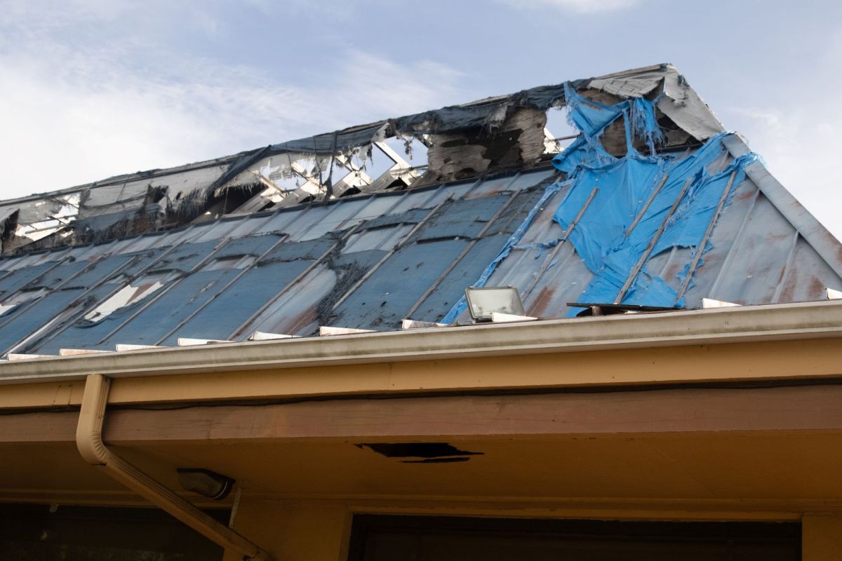 A damaged roof covered with blue tarps and exposed areas under a partly cloudy sky.