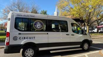 An electric shuttle bus parked on a street with trees and a church steeple in the background.