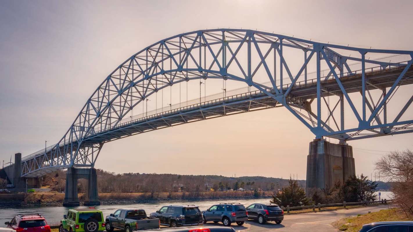 The large arched Sagamore Bridge spans over a river with parked vehicles visible along the shore.