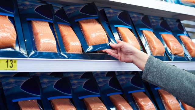 A hand reaches for a package of salmon fillets on a supermarket shelf.